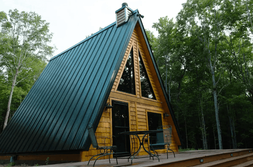 A-frame cabin with a green roof and wooden deck surrounded by trees