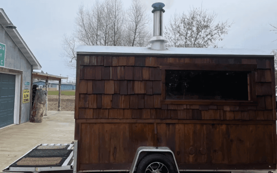 Wood-shingled sauna trailer with a chimney beside a building and bare trees