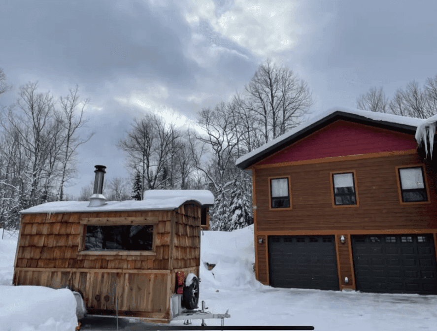 Wood-shingled sauna trailer beside a house in deep snow