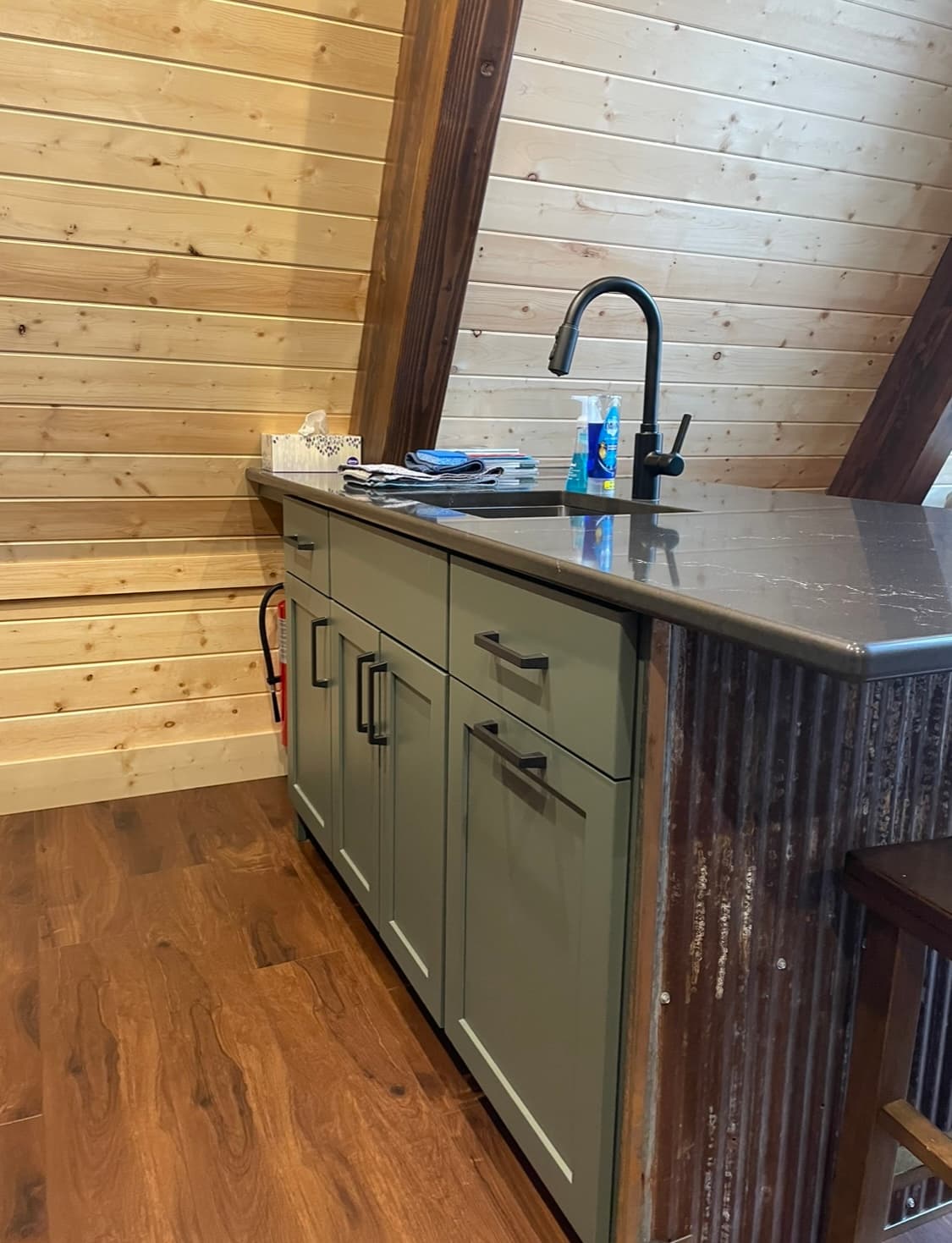 Cabin kitchen sink and green lower cabinets beneath sloping wood walls