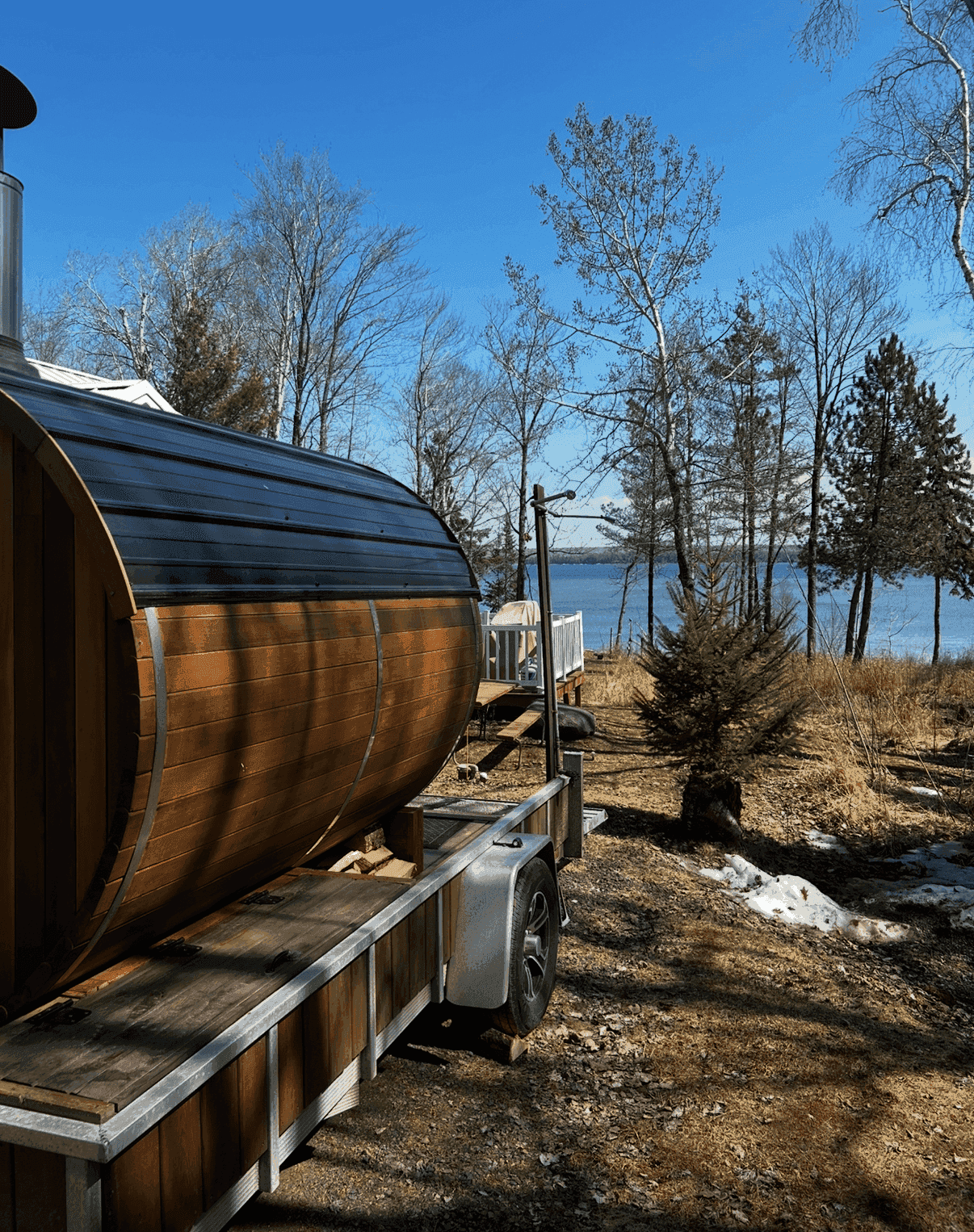 Barrel sauna trailer beside a lake and bare trees